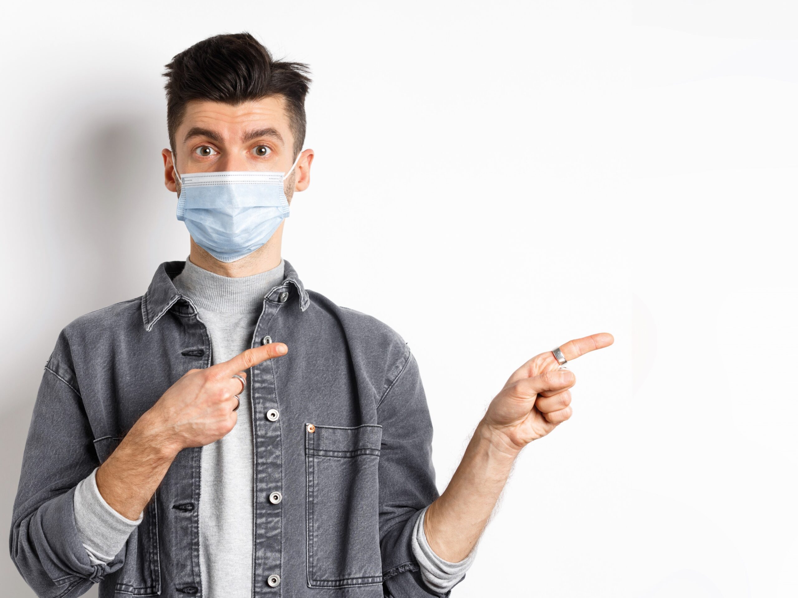 Pandemic lifestyle, healthcare and medicine concept. Young guy in face mask inviting look here, pointing fingers left at logo, standing on white background